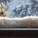 A pristine white Arctic Fox Pelt, soft and luxurious, draped elegantly over a wooden trader’s table. The background features a rustic northern outpost with snow-covered mountains visible through an open window, and other furs and pelts stacked neatly for trade. The lighting is soft and cool, emphasizing the fur’s warmth and delicate texture, evoking a sense of arctic elegance and rugged beauty